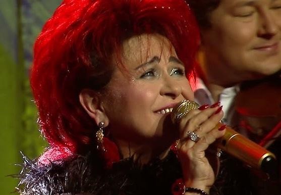 Bernadeta Kowalska na scenie podczas koncertu, śpiewająca do mikrofonu, w otoczeniu świateł scenicznych, z radosnym wyrazem twarzy.