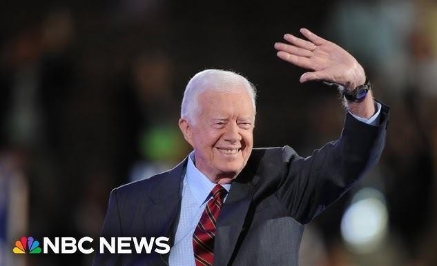 Jimmy Carter w ubraniu roboczym aktywnie budujący dom w ramach Habitat for Humanity, z uśmiechem na twarzy, w otoczeniu innych wolontariuszy i częściowo zbudowanego domu.