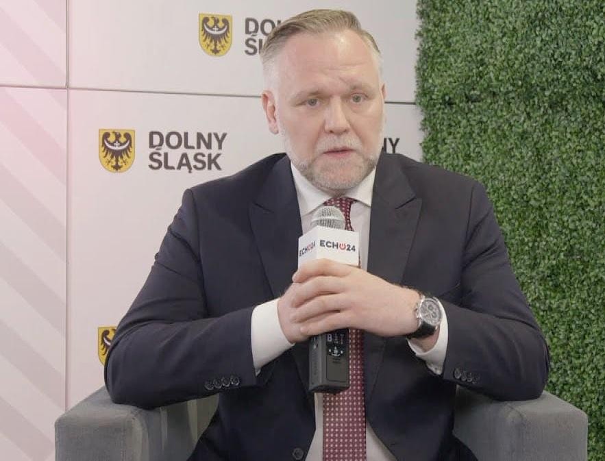 Dawid Jackiewicz przemawiający na spotkaniu, gestykulujący, w tle zamazany ekran prezentacyjny.