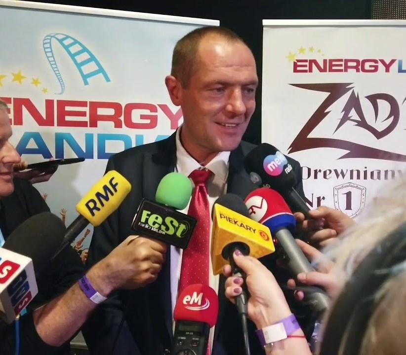Marek Goczał w kombinezonie rajdowym, stojący obok samochodu rajdowego SSV na tle pustynnego krajobrazu, symbolizujący jego pasję do rajdów terenowych.